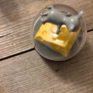 May include: A clear plastic spherical container enclosing a small gray mouse figurine positioned above a yellow block of Swiss cheese with visible holes. The mouse is sculpted in a dynamic pose, and the entire assembly is contained within the transparent dome.