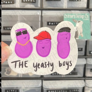 May include: A white sticker with three purple cartoon characters wearing hats and sunglasses. The characters are labeled "THE yeasty boys".