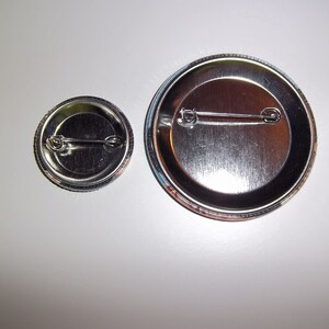 Peut inclure: Deux badges m&eacute;talliques de tailles diff&eacute;rentes, vus de dos. Le plus grand badge a un diam&egrave;tre d'environ 7,6 cm, tandis que le plus petit mesure environ 3,8 cm. Les deux ont une finition argent&eacute;e brillante et une attache de type &eacute;pingle de s&ucirc;ret&eacute;.