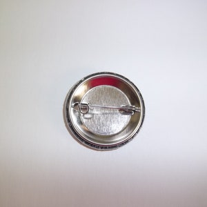 Peut inclure: Un bouton de badge argent&eacute; avec une fermeture &agrave; &eacute;pingle de s&ucirc;ret&eacute;. Le dos du bouton est visible, montrant la construction m&eacute;tallique et le m&eacute;canisme de l'&eacute;pingle. Le bouton a une surface brillante et r&eacute;fl&eacute;chissante.