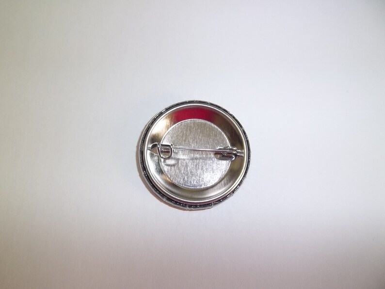 Peut inclure: Un bouton de badge argent&eacute; avec une fermeture &agrave; &eacute;pingle de s&ucirc;ret&eacute;. Le dos du bouton est visible, montrant la construction m&eacute;tallique et le m&eacute;canisme de l'&eacute;pingle. Le bouton a une surface brillante et r&eacute;fl&eacute;chissante.