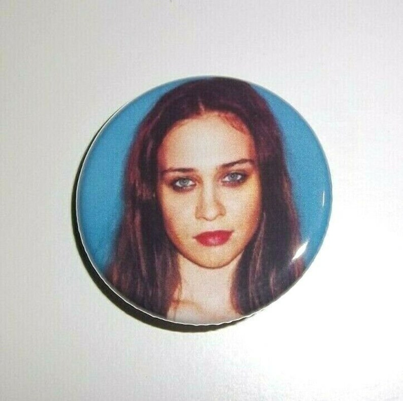 Peut inclure: Un bouton rond avec un portrait de femme aux cheveux bruns, aux yeux bleus et au rouge &agrave; l&egrave;vres rouge, sur fond bleu clair. Un objet de collection.