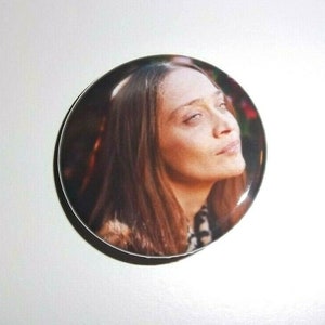 Peut inclure: Un bouton rond avec un portrait d'une femme aux longs cheveux bruns. L'image est un gros plan de son visage, avec un l&eacute;ger regard vers le haut. Le bouton a une finition brillante et un bord blanc.