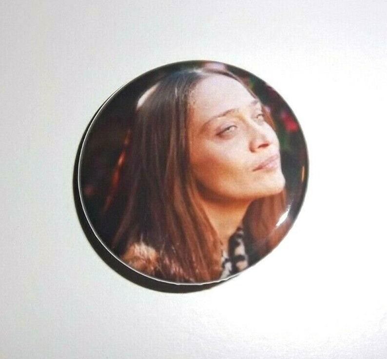 Peut inclure: Un bouton rond avec un portrait d'une femme aux longs cheveux bruns. L'image est un gros plan de son visage, avec un l&eacute;ger regard vers le haut. Le bouton a une finition brillante et un bord blanc.