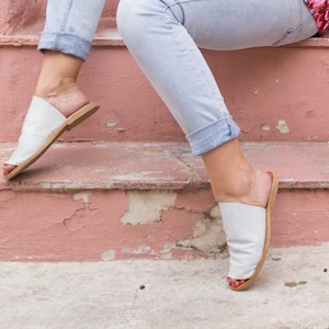 May include: A pair of white leather sandals with a brown sole. The sandals are worn by a person sitting on a set of pink stairs.
