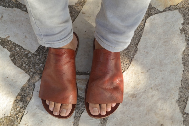 Puede incluir: Un par de sandalias de cuero marr&oacute;n con un dise&ntilde;o sencillo. Las sandalias son usadas por una persona con los pies descalzos. Las sandalias est&aacute;n sobre un suelo de piedra.