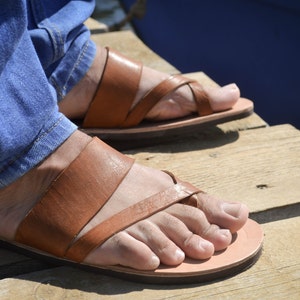 Pu&ograve; includere: Un paio di sandali in pelle marrone con un design semplice. I sandali hanno un cinturino che attraversa il piede e un cinturino per le dita. I sandali sono indossati da una persona che indossa jeans blu.
