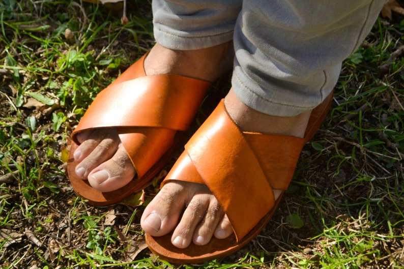 May include: A pair of brown leather sandals with a criss-cross strap design. The sandals are worn on a person's feet and are set against a background of green grass.