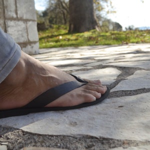 May include: A person's foot wearing a black leather thong sandal on a stone pathway.