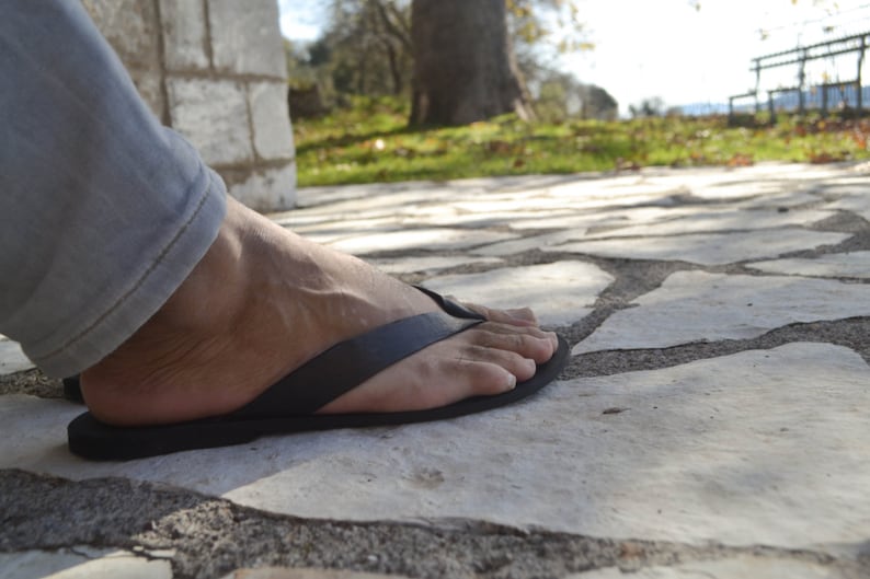 May include: A person's foot wearing a black leather thong sandal on a stone pathway.