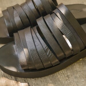 Puede incluir: Un par de sandalias de cuero negro con un dise&ntilde;o de tiras tejidas. Las sandalias tienen una suela gruesa y plana.
