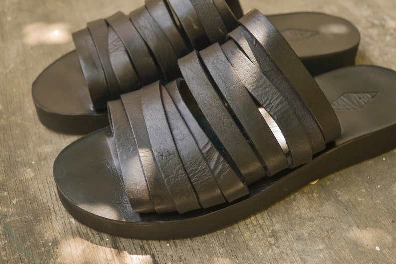 Puede incluir: Un par de sandalias de cuero negro con un dise&ntilde;o de tiras tejidas. Las sandalias tienen una suela gruesa y plana.