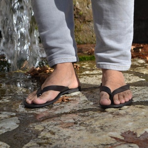 May include: A person wearing grey jeans and black flip-flops standing on a stone path near a waterfall.
