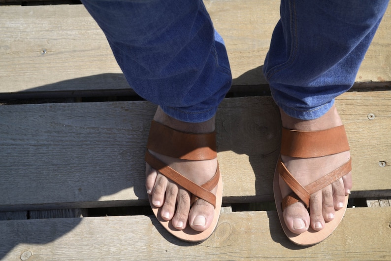 Pu&ograve; includere: Un paio di sandali in pelle marrone con un design a cinturino incrociato. I sandali sono indossati su una superficie di legno.
