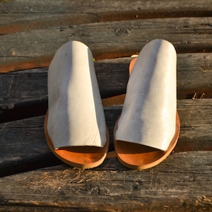 May include: A pair of white leather sandals with brown leather soles. The sandals are on a wooden surface.