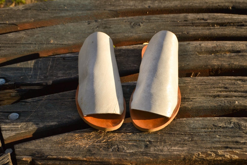 May include: A pair of white leather sandals with brown leather soles. The sandals are on a wooden surface.