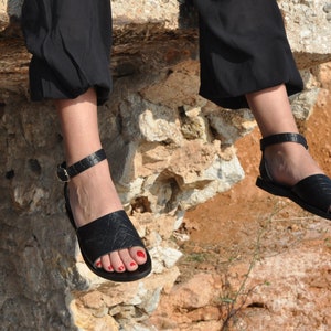 May include: Black leather sandals with ankle straps and gold buckles. The sandals have a textured design on the straps and footbed. The image shows the sandals on a person's feet, with red nail polish on the toes.