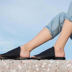 May include: A pair of black leather slide sandals with a simple design. The sandals are worn by a person sitting on a concrete surface.