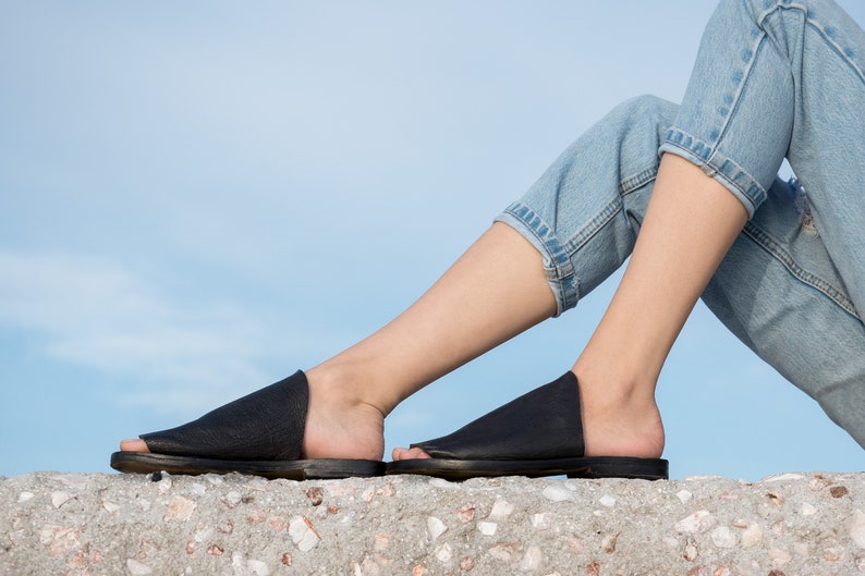 May include: A pair of black leather slide sandals with a simple design. The sandals are worn by a person sitting on a concrete surface.
