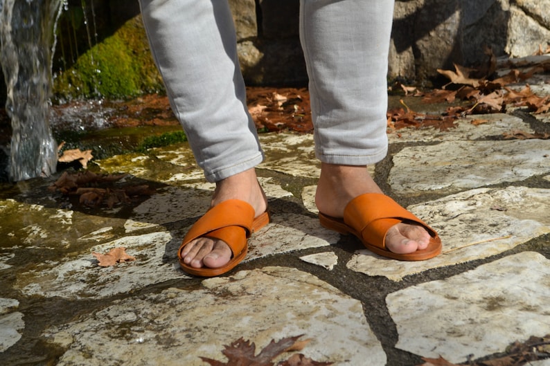 May include: A pair of brown leather sandals with a criss-cross strap design. The sandals are worn by a person standing on a stone path.