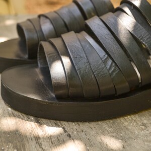 Puede incluir: Un par de sandalias de cuero negro con suela plana y m&uacute;ltiples tiras. Las sandalias est&aacute;n hechas de cuero suave y flexible y tienen un aspecto informal y c&oacute;modo.