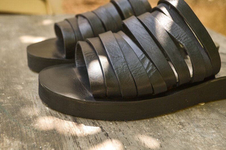 Puede incluir: Un par de sandalias de cuero negro con suela plana y m&uacute;ltiples tiras. Las sandalias est&aacute;n hechas de cuero suave y flexible y tienen un aspecto informal y c&oacute;modo.