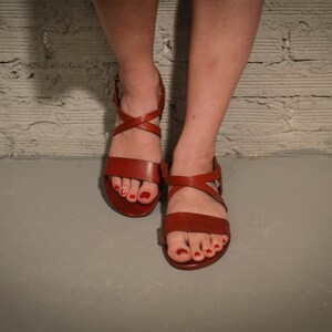 May include: A pair of brown leather sandals with criss-cross straps and buckles. The sandals have red nail polish on the toes.