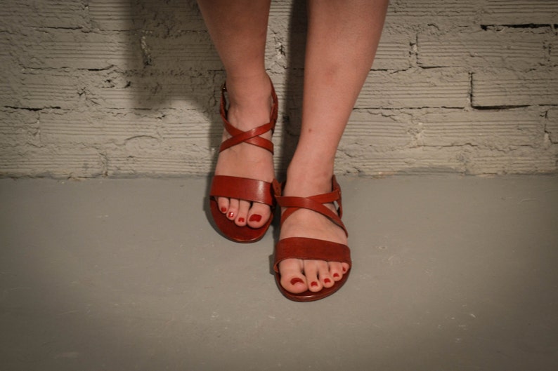 May include: A pair of brown leather sandals with criss-cross straps and buckles. The sandals have red nail polish on the toes.