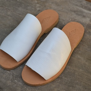 May include: A pair of white leather sandals with a brown leather sole. The sandals have a simple design and are perfect for summer.