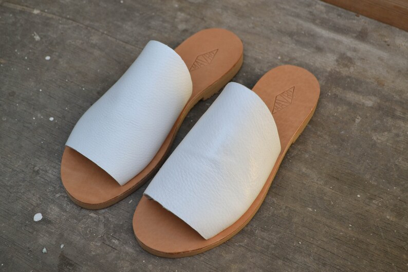 May include: A pair of white leather sandals with a brown leather sole. The sandals have a simple design and are perfect for summer.