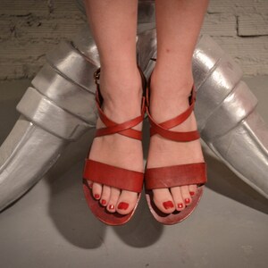 May include: A pair of red leather sandals with criss-cross straps and a buckle closure. The sandals have a low heel and are worn on a person's feet.