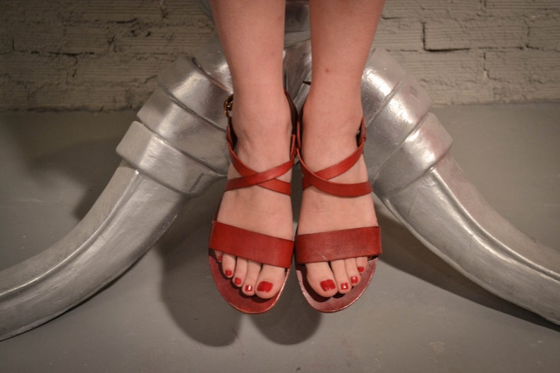 May include: A pair of red leather sandals with criss-cross straps and a buckle closure. The sandals have a low heel and are worn on a person's feet.