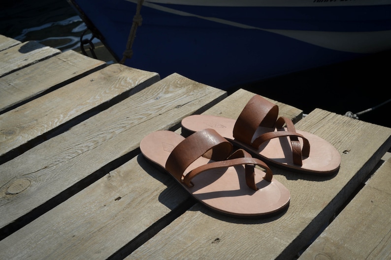 Pu&ograve; includere: Due paia di sandali in pelle marrone con cinturini. I sandali sono su una superficie di legno.