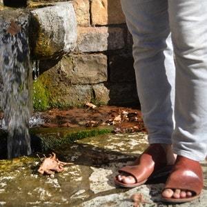 Puede incluir: Una persona con jeans de color claro y sandalias de cuero marr&oacute;n se encuentra cerca de una fuente de piedra con agua fluyendo.