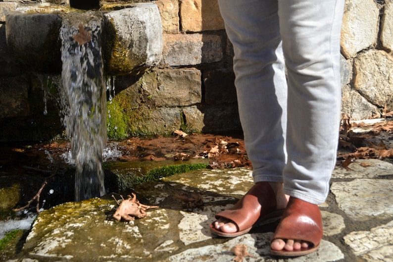Puede incluir: Una persona con jeans de color claro y sandalias de cuero marr&oacute;n se encuentra cerca de una fuente de piedra con agua fluyendo.