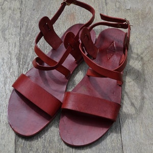 May include: A pair of red leather sandals with multiple straps and buckles. The sandals have a simple design and are made of high-quality leather.