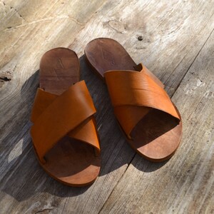 May include: A pair of brown leather sandals with a criss-cross strap design. The sandals are on a wooden surface.