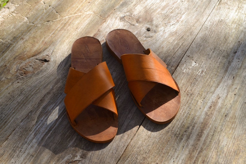 May include: A pair of brown leather sandals with a criss-cross strap design. The sandals are on a wooden surface.