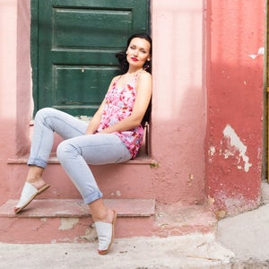 May include: A woman wearing a floral print tank top and light blue jeans sits on a pink and red painted step. She is wearing white sandals and has her hair pulled back.
