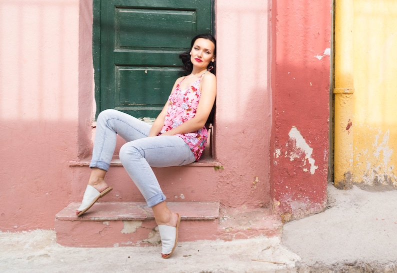 May include: A woman wearing a floral print tank top and light blue jeans sits on a pink and red painted step. She is wearing white sandals and has her hair pulled back.