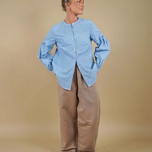 Op de afbeelding: Een lichtblauw overhemd met een mandarijnkraag en volumineuze lange mouwen met geplooide manchetten, gecombineerd met een losvallende beige broek en donkerbruine teenslippers. Het overhemd heeft een borstzak en een knoopsluiting aan de voorzijde.