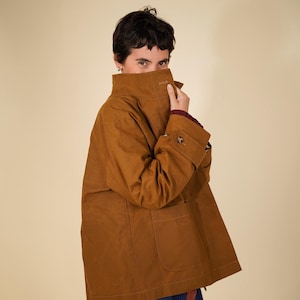 May include: A tobacco brown jacket featuring a high collar, long sleeves with buttoned cuffs, and two large front patch pockets. The jacket is constructed with visible stitching and is worn over a dark blue pleated skirt and a maroon sweater.