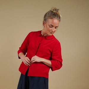 Pode incluir: Uma camisa vermelha brilhante com botões, gola arredondada e mangas três quartos, combinada com uma saia longa e fluida azul-marinho. O visual é completado com ténis baixos castanhos e brancos com um design desportivo clássico.