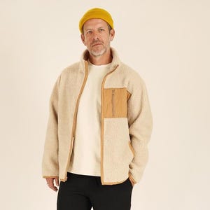 May include: A person wearing a beige fleece jacket with a brown pocket and trim, a white t-shirt, black pants, a yellow beanie, and beige and red sneakers. The jacket has a high collar and a zipper closure.