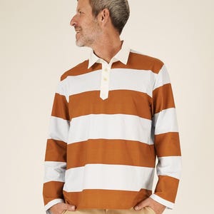 May include: A long-sleeved rugby shirt with horizontal stripes in brown, white, and light blue. The shirt has a white collar and a three-button placket. The person is wearing khaki pants.