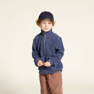 Puede incluir: Un niño pequeño con pelo rojo que lleva una chaqueta azul con cremallera, una gorra de béisbol negra y pantalones de cuadros marrones.