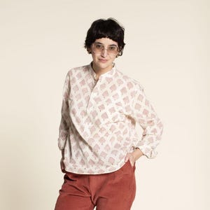 May include: A white long-sleeved shirt with a pink floral print. The shirt has a mandarin collar and a button closure. The model is wearing the shirt with red pants.