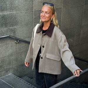 May include: A beige jacket featuring a dark brown corduroy collar, long sleeves, and a front button closure. The garment includes a single flap pocket on the lower left side and is worn over a black shirt paired with dark gray denim jeans.