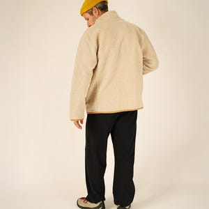 May include: A person wearing a yellow beanie, a beige fleece jacket with brown trim, black pants, and tan and red shoes. The person is facing away from the camera.