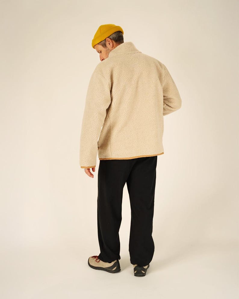 May include: A person wearing a yellow beanie, a beige fleece jacket with brown trim, black pants, and tan and red shoes. The person is facing away from the camera.